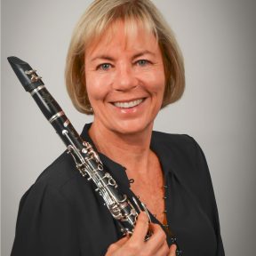 Susan Macy poses with a smile with her clarinet in front of a plain backdrop.