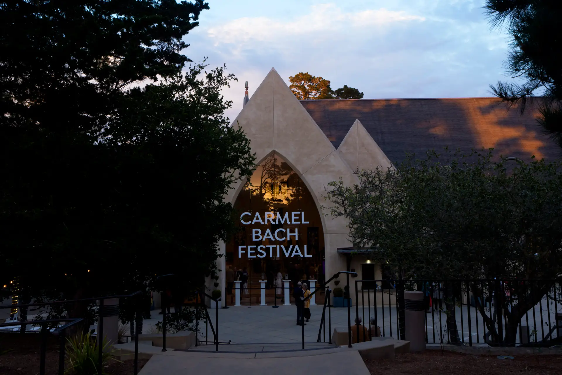 The lit entrance arch of Sunset Center at dusk, with the Carmel Bach Festival name across the glass and patrons gathering on the plaza.