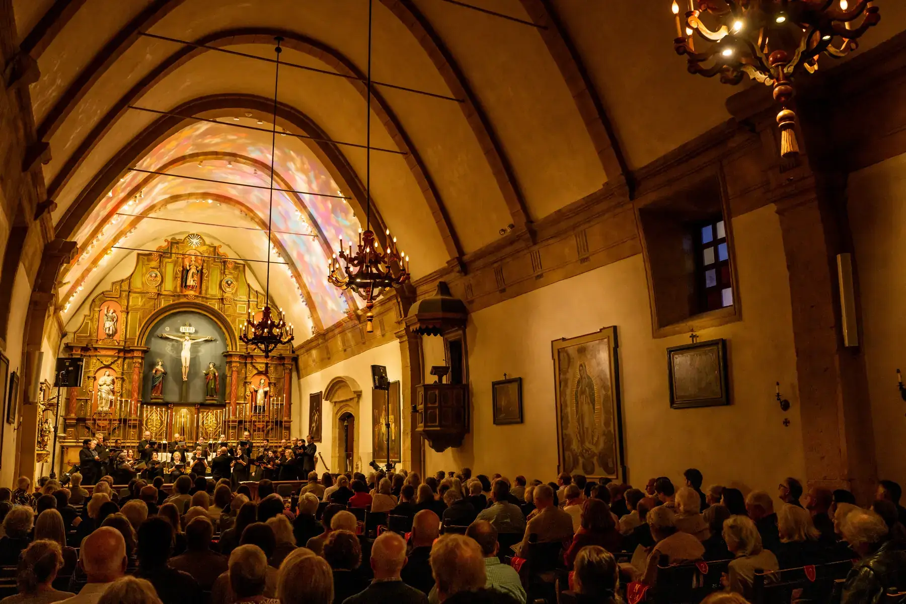 A choir performs before the gilded altar of the Carmel Mission Basilica beneath its arched ceiling and candle chandeliers, with a full audience in the pews.