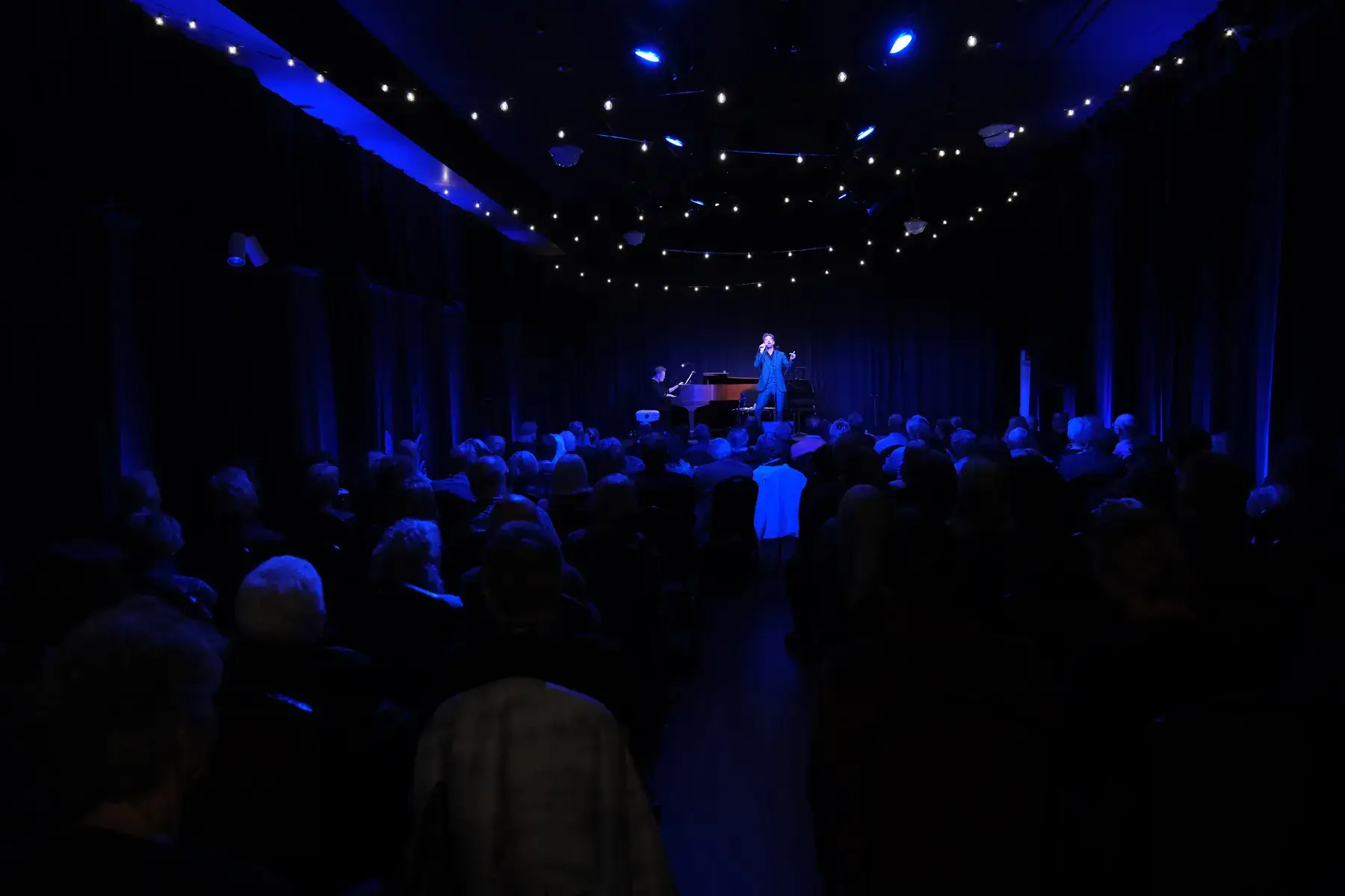 An audience seated in Studio 105 under strings of café lights, watching a singer and pianist perform in blue stage light.