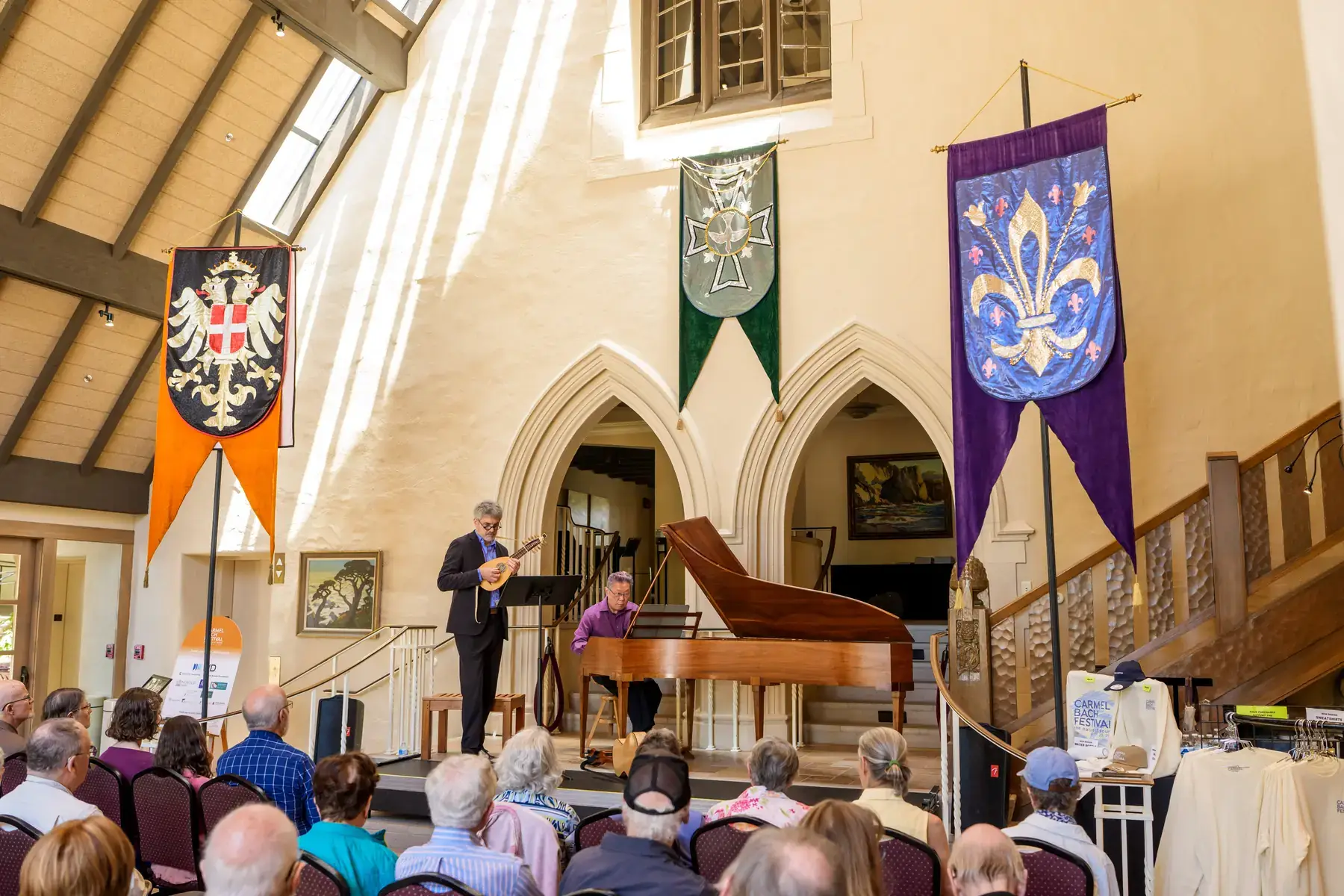 A lutenist and harpsichordist perform in the sunlit Sunset Center Foyer beneath tall heraldic banners, with an audience seated in front.
