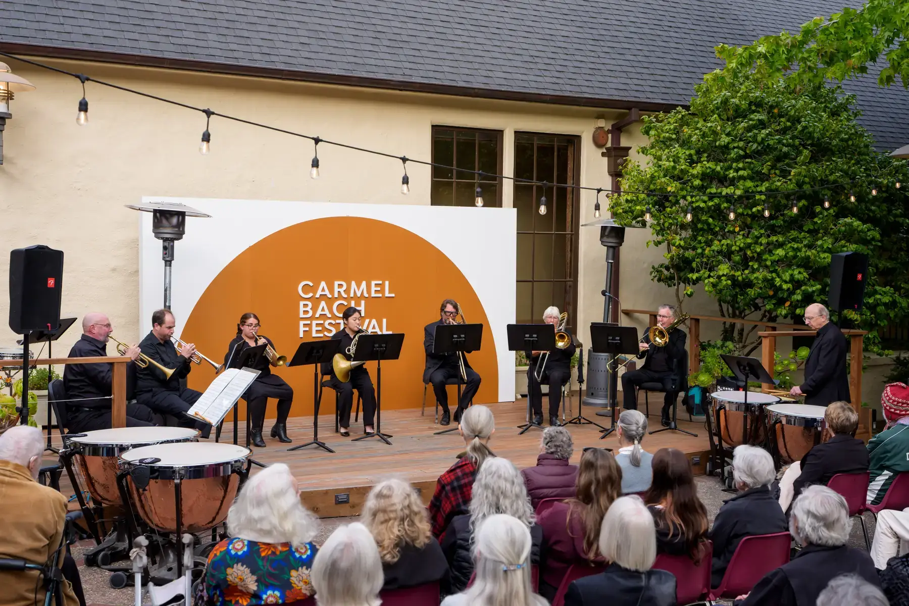 A brass and timpani ensemble performs on the Sunset Terrace stage under string lights, in front of a Carmel Bach Festival backdrop.