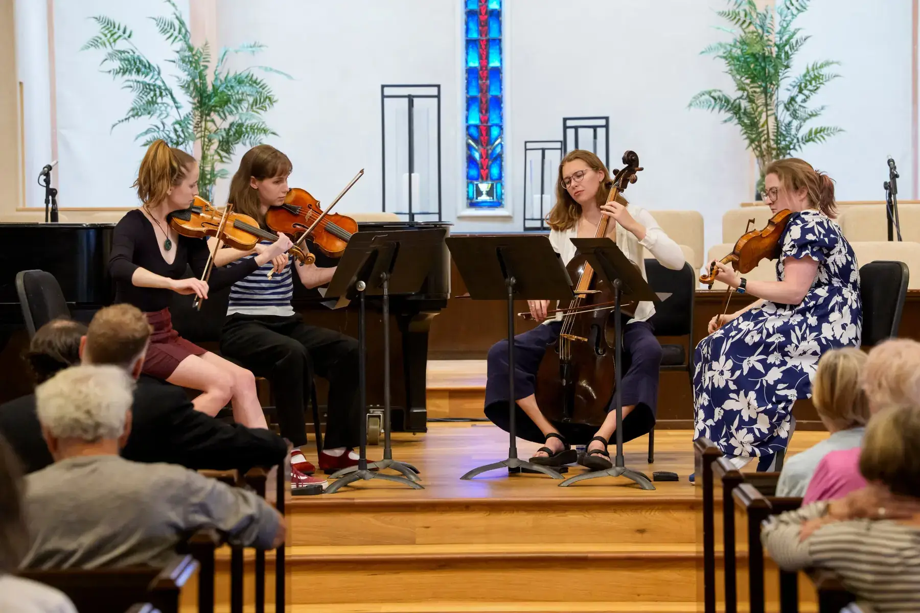 A string quartet performs on the chancel steps at Carmel Presbyterian Church, beneath a tall blue stained-glass window, for an audience in the pews.